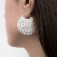 Large silver disc hoop style earrings with textured finish on model.